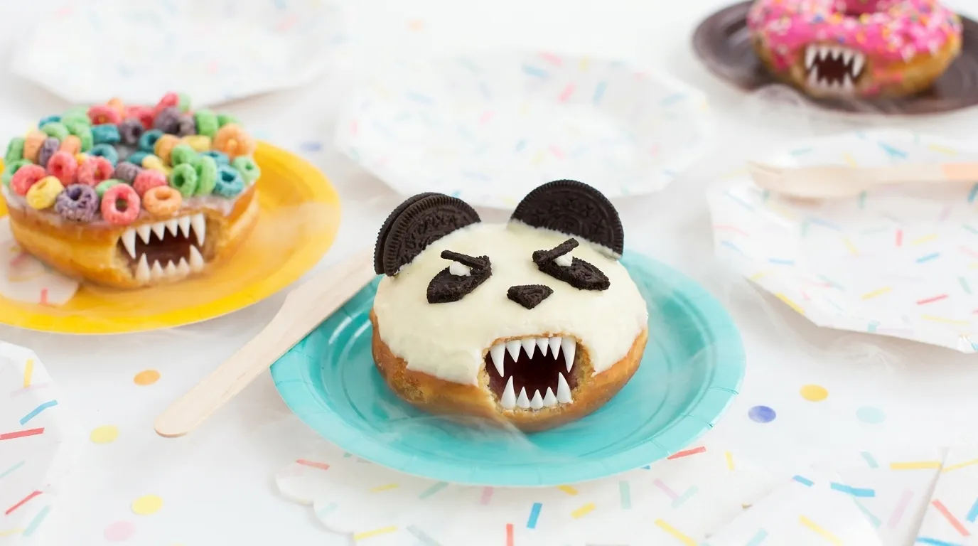A festive table of colorful party donuts on paper plates with sprinkle confetti scattered around, now horrifyingly alive: each donut has sprouted a wide grinning mouth full of sharp fangs, its candy-eyes narrowed into a menacing glare, bright party lighting and thin mist crawling across the tabletop
